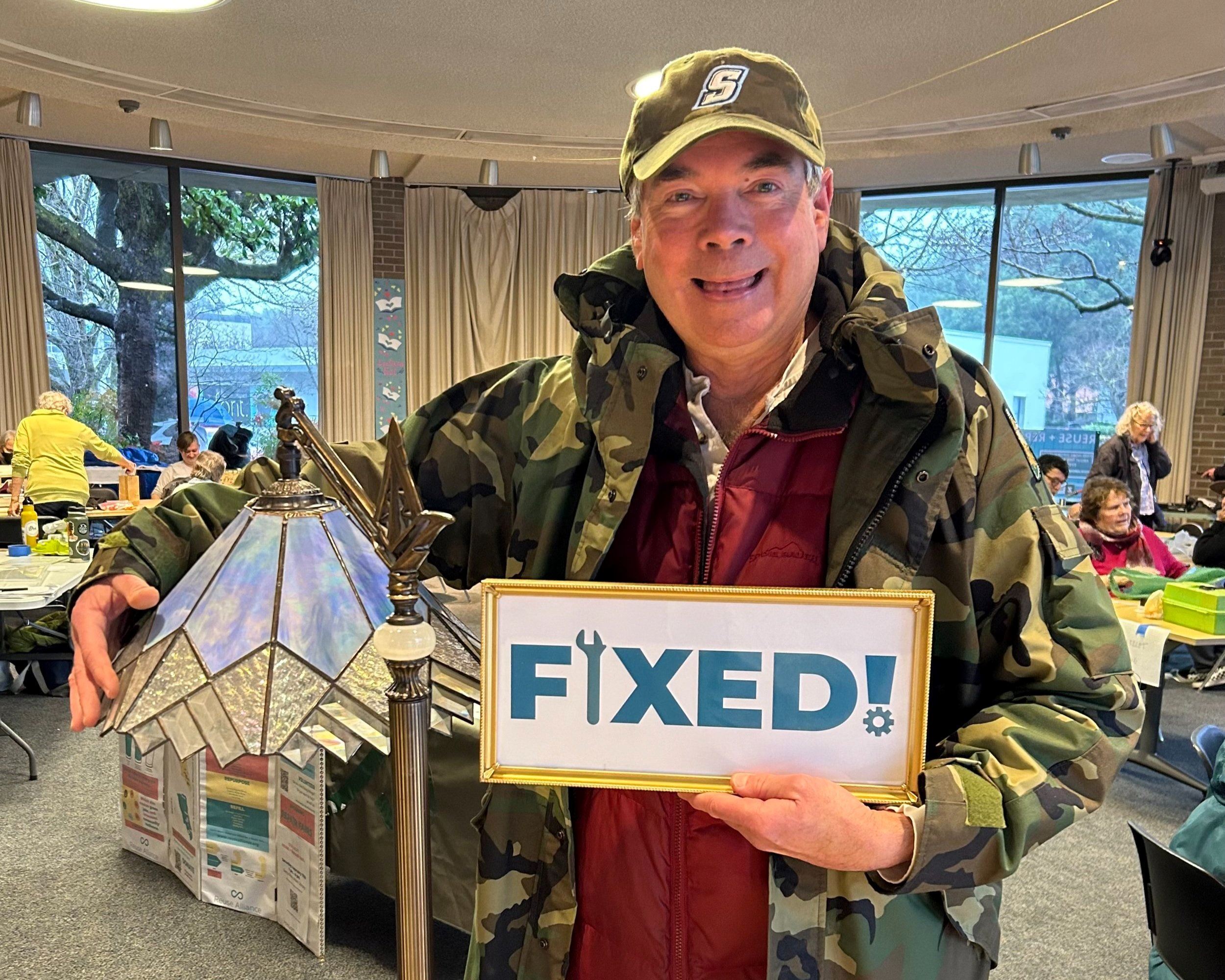 A smiling man holds a Tiffany-style stained glass lamp in one hand and a sign that says fixed