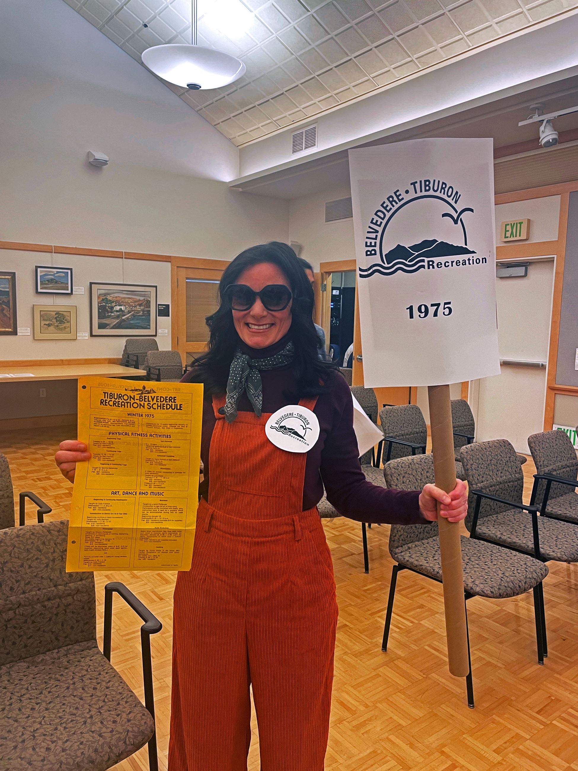 A brunette woman dressed in 1970s clothing holding a sign that says Belvedere-Tiburon Recreation